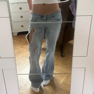 Ljusblå bootcut jeans med slitningar - Säljer ett par ljusblå bootcut jeans med snygga slitningar och hål på benen. Jeansen har låg midja och klassisk femficksmodell. Perfekta för en avslappnad och trendig look. Materialet är jeans i bomull. Inmerbensläng: 81cm, midjemått: 42cm