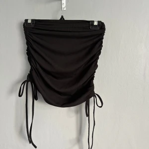 Svart ribbad kortkjol med snörning - Supersnygg svart kortkjol med ribbad struktur och trendig snörning i sidorna. Kjolen har rynkade detaljer längs sidorna som ger en cool och figurnära passform. Perfekt för dig som gillar en stilren men edgy look.