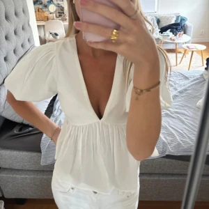 Vit blus med puffärm och v-ringning - Supersöt vit blus med djup v-ringning och puffiga korta ärmar. Modellen har en lätt rynkad midja som ger en flowy känsla och en feminin siluett. Perfekt för dig som gillar romantiska och trendiga detaljer.