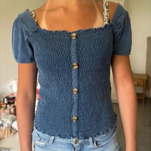 Blå smockad blus från Zara - Supersöt blå smockad blus från Zara med kort puffärm och snygga träknappar framtill. Storlek 13-14 år men den är stor så storlek XS-S