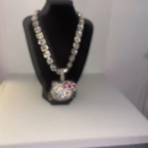 Glittrigt Hello Kitty-halsband - Ett statement halsband med en stor Hello Kitty-berlock täckt av glittrande stenar. Kedjan är silverfärgad och består av fyrkantiga länkar med infattade stenar. Hello Kitty-berlocken har rosa och röda detaljer på rosetten och gnistrar rejält.