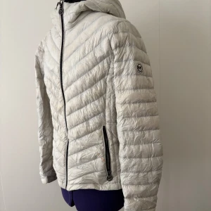 Ljusgrå dunjacka från Michael Kors - Snygg ljusgrå dunjacka från Michael Kors med huva och dragkedja framtill. Jackan har quiltad design i sicksackmönster och två fickor med dragkedja. Perfekt för kyliga dagar och enkel att packa ner när du är på språng.