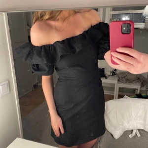 Svart offshoulder kortklänning - Supersnygg svart kortklänning med offshoulder och volang upptill. Klänningen har puffiga korta ärmar och en figurnära passform som framhäver siluetten. Perfekt för dig som vill sticka ut med en trendig och elegant look.
