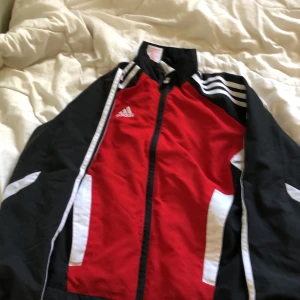 Adidas röd och svart vindjacka - Säljer en sportig vindjacka från Adidas i rött, svart och vitt. Jackan har dragkedja framtill, ståkrage och klassiska vita ränder på axlarna. Perfekt för dig som gillar en aktiv stil och vill sticka ut med färg. Adidas-logga på bröstet och praktiska fickor på sidorna.