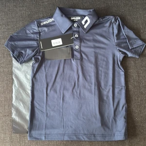 Mörkblå pikétröja från Druids 6/8 years - Snygg mörkblå pikétröja från Druids med klassisk krage och tre knappar framtill. Tröjan har korta ärmar och logotyp på både krage och axel. Perfekt för en sportig och stilren look, tillverkad i mjukt material som känns skönt mot huden. Säljs då jag fick fel storlek och returfrakten var 200:-, bättre någon får köpa den istället för att skickas tillbaka.