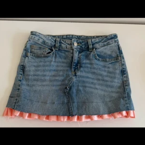 Jeanskjol med rosa volangkant - Unik kort jeanskjol med rå kant längst ner, som jag sytt om från ett par jeans. Kjolen har en volang i glansig rosa satin som sticker ut under fållen och ger en cool kontrast till den klassiska denimlooken. Perfekt för dig som vill sticka ut lite extra!
