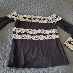 Svart Intimissimi offshoulder topp med spetsdetaljer - Supersnygg svart Intimissimi  offshoulder topp med långa ärmar och breda, vita spetsband med blommönster över bröst, ärmslut och axlar. Toppens romantiska vibe gör den till ett statement-plagg. Perfekt för dig som gillar unika detaljer och vill sticka ut. Intimissimi lapp är klippt. Storlek S. Den är I väldigt fin skick