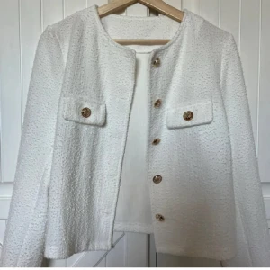 Vit cardigan jacka med guldknappar - Stilren vit cardigan jacka med snygga guldknappar framtill och på fickorna. Jackan har rak passform, rund halsringning och är tillverkad i ett strukturerat tyg som ger en exklusiv känsla. Perfekt att slänga över en topp för en chic look.