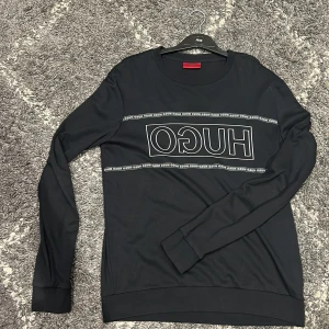 Svart långärmad tröja från Hugo Boss - Svart långärmad tröja från Hugo Boss med stor vit logga framtill. Tröjan har rund halsringning och är tillverkad i mjukt material som känns skönt mot huden. Perfekt för dig som gillar stilren och enkel design med tydlig branding. Knappt använd!
