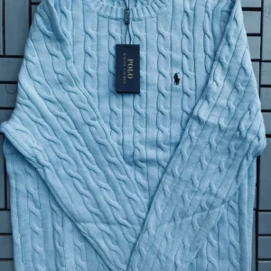 Ljusblå stickad tröja från Polo Ralph Lauren - Säljer en stilren ljusblå stickad tröja från Polo Ralph Lauren med det klassiska broderade logotypen på bröstet. Tröjan har en kabelstickad design och rund halsringning. Perfekt för kyligare dagar.