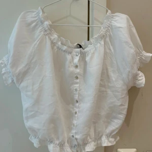 Vit blus med knappar och resår - Supersöt vit blus från Zara  med korta puffärmar och resår vid hals, ärmslut och midja. Blusen har knappar framtill och är gjord i ett lätt och luftigt linnematerial, perfekt för varma dagar. Enkel och fräsch stil som passar till jeans eller kjol.