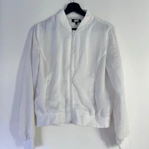 Vit bomberjacka från Bik Bok - Snygg vit bomberjacka från Bik Bok med dragkedja framtill och ribbade muddar vid ärmslut och midja. Jackan har två fickor med dragkedja och klassisk bomberkrage. Perfekt för dig som gillar en clean och stilren look.