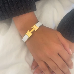 Hermèns vit och guld Clic H armband - Säljer ett Hermèns Clic H armband i vit emalj med guldfärgade metalldetaljer. Armbandet har den klassiska H-formade låset och en stilren, elegant design som verkligen sticker ut. Perfekt accessoar för att lyfta vilken outfit som helst.