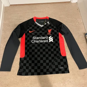 Liverpool långärmad bortatröja Nike S - Liverpool FC långärmad bortatröja från Nike i svart och grått schackrutigt mönster. Röda detaljer på sidorna, kragen och loggan. Tillverkad i lätt och ventilerande Dri-FIT-material. V-ringad hals och tryckt sponsor på bröstet.