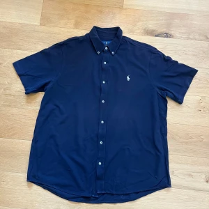 Mörkblå skjorta från Ralph Lauren - Snygg mörkblå kortärmad skjorta från Ralph Lauren i storlek XXL. Skjortan har klassisk button-down krage, vita knappar och den ikoniska broderade polospelaren på bröstet. Tillverkad i lätt och luftig bomullsmix, perfekt för varma dagar. Ställ gärna frågor!