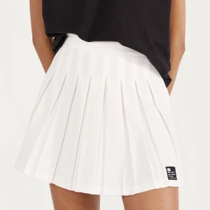 Vit plisserad kjol från Bershka - Superfin vit kort plisserad kjol från Bershka. Kjolen har bred linning och tydliga veck som ger en snygg och sportig look. Perfekt att matcha med en crop top eller hoodie för en trendig stil. Liten svart logga nertill på ena sidan.