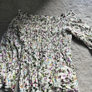 Blommig offshoulder blus från Ellos - Supersöt blommig blus från Ellos i vitt med färgglada blommor. Offshoulder-modell med smock upptill och långa ärmar. 