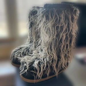 Päls ”moon boots” - Unika boots med lång, lurvig pälsimitation i svart och grått. Skorna har en platt sula och rund tå, perfekt för dig som vill sticka ut med en cool och annorlunda stil. 