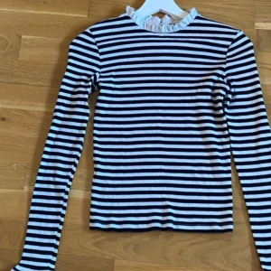 Randig topp från Mango! Nyskick! -  Säljer en stilren randig topp i svart och vit med långa ärmar. Toppen har en söt volangdetalj vid halsen och en knappöppning i nacken. Perfekt för en chic och avslappnad look. Är i jättefint skick! Från axel t axel 35 cm  tröjans total längd 52 cm. Den är stretchig. 