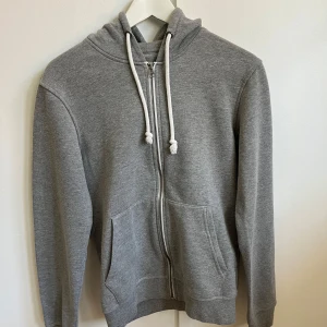 Grå hoodie med dragkedja från Pier One - Klassisk grå hoodie från Pier One med vit snörning i huvan och hel dragkedja framtill. Tröjan har två stora fickor och är tillverkad i mjuk bomullsmix.