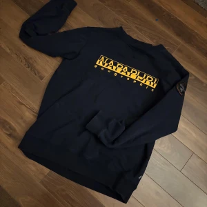 tröja från Napapijri - Säljer en svart tröja från Napapijri med gult tryck på bröstet och en snygg patch på ärmen. Tröjan har långa ärmar och ribbade muddar vid hals och ärmslut. Perfekt för en stilren look. Storlek M men ska nog passa S också.