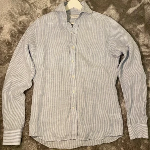 Randig linne skjorta från John Henric - Snygg blå- och vitrandig skjorta från John Henric. Skjortan har långa ärmar och knappar framtill. Perfekt för en stilren look. Storleken på tröjan är ungefär S. Har bara använt den några gånger men har växt ur den.