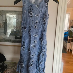 Blommig blå klänning från Alesia International - Säljer en blå klänning med blommigt mönster från Alesia International. Klänningen har en dragkedja i ryggen och snörning bak över dragkedjan för en justerbar passform. Perfekt för sommarens alla tillfällen!