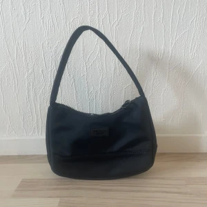 Svart axelväska med dragkedja - Snygg svart axelväska i syntetmaterial med praktisk dragkedja. Perfekt för att bära dina nödvändigheter med stil. Enkel och elegant design som passar till många tillfällen.