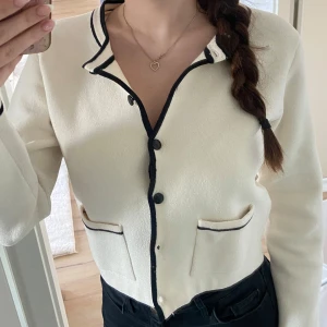 Vit kofta från Reserved - Säljer en stilren vit kofta från Reserved med svarta detaljer. Koftan har en v-ringad design och knappar framtill. Den är långärmad och har fickor med svarta kanter. Perfekt för en elegant look.