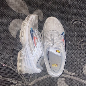 Vita Nike sneakers med färgglada detaljer - Säljer ett par vita Nike sneakers med synlig luftdämpning och färgglada detaljer i blått, rött och orange. Skorna har snörning och en sportig design, perfekt för en casual look.