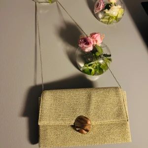  kuvertväska - Elegant beige kuvertväska med en stilren kedja som axelrem. Väskan har en rund guldfärgad knapp som stängning och är perfekt för att bära det nödvändigaste. Den har en enkel och chic design som passar till många tillfällen. Använd 2 ggr, inga detekter. 🌼🥂☀️