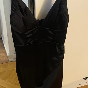 Svart sidenklänning - Elegant svart klänning i siden med djup v-ringning. Perfekt för en kväll ute eller en speciell tillställning. Klänningen har en smickrande passform och glänsande yta som ger en lyxig känsla.