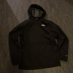 Svart regnjacka från The North Face - Säljer en stilren svart regnjacka från The North Face. Jackan har en praktisk huva och dragkedja framtill. Perfekt för regniga dagar och håller dig torr och bekväm. The North Face-loggan syns både fram och bak. Det är storlek xxl i barn men passar s