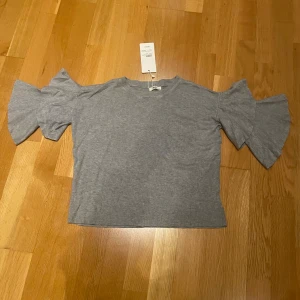 Grå topp med volangärmar från Missäy - Säljer en snygg grå topp från Missäy med volangärmar och offshoulder-design. Aldrig använd pågrund av att jag inte hann lämna tillbaka den. Perfekt för att ge en trendig touch till din outfit. Toppen är i en bekväm passform och tillverkad i mjukt material.
