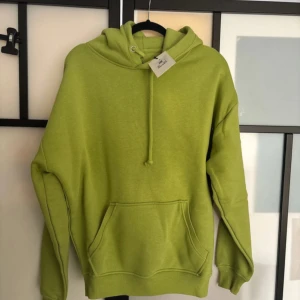 Grön hoodie med ficka - Säljer en fräsch grön hoodie med en stor ficka framtill och justerbar huva. Perfekt för en avslappnad stil. Den har en mjuk och bekväm passform.