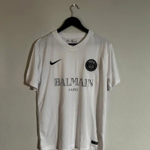 PSG x Balmain Kit - Snygg vit PSG x Balmain fotbollströja från Nike. Tröjan har korta ärmar och är tillverkad i Dri-FIT-material för optimal komfort. Den har PSG-loggan och Balmain Paris-tryck på framsidan. Perfekt för fotbollsfans som vill ha stil på planen. Nyskick!