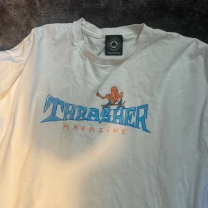  Thrasher T-shirt  - Säljer en vit t-shirt från Thrasher  med ett coolt tryck i blått och orange på framsidan. T-shirten har en klassisk passform och är kortärmad. Perfekt för en avslappnad stil! 