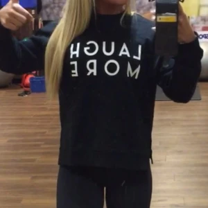 Svart sweatshirt med text - Säljer en super mysig svart sweatshirt med texten 'LAUGH MORE' i vitt på framsidan. Tröjan har långa ärmar och en bekväm passform, med slits på sidorna. 