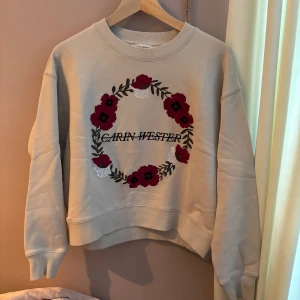 Beige sweatshirt Carin Wester - Säljer en beige sweatshirt från Carin Wester med ett broderat blommönster i rött och vitt på framsidan. Tröjan har långa ärmar och en rund halsringning. Nästan helt oanvänd