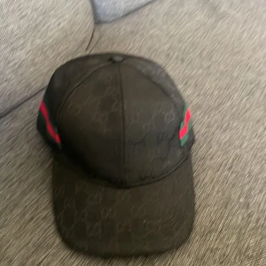Svart keps från Gucci - Snygg svart keps från Gucci med diskret mönster och röd-gröna detaljer på sidorna. Perfekt för att ge din outfit en lyxig touch.