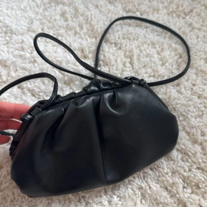 Dumpling bag bow 19 - Snygg svart axelväska i syntetmaterial med en stilren design. Väskan har en enkel axelrem och en rymlig öppning för enkel åtkomst. Perfekt för att bära med sig det nödvändigaste. Helt ny, aldrig använd