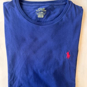 Blålila t-shirt från Ralph Lauren - Säljer en blå t-shirt från Ralph Lauren med röd broderad logga på bröstet. Tröjan satt som en smäck tills det att jag blev lite större - vilket var mycket synd, för det var inte många gånger jag hann använda den. En tisha som är perfekt för en skön, avslappnad och snygg look helt enkelt. 
