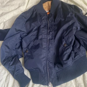 Mörkblå bomberjacka från Alpha Industries - Säljer en snygg mörkblå bomberjacka från Alpha Industries. Jackan har en dragkedja framtill och ribbade muddar vid ärmslut och nederkant. Perfekt för en cool och stockholmsstil look. 