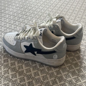 Gråvita sneakers från BAPE - Snygga BAPE sneakers i grått och vitt med den ikoniska stjärndetaljen i svart på sidan. nästan inga tecken på användning då jag köpte för ungefär en månad sedan. Jag säljer dom för att dem är för små. 