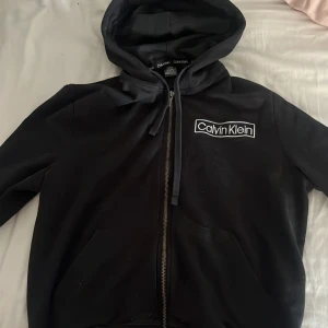 Svart hoodie från Calvin Klein - Säljer en stilren svart hoodie från Calvin Klein med dragkedja och logga på bröstet. Perfekt för en avslappnad look. Den har en praktisk huva och fickor framtill.