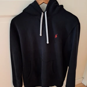 Svart hoodie från Ralph Lauren - Säljer en klassisk svart hoodie från Ralph Lauren med vit dragsko och det ikoniska röda logotypen på bröstet. Perfekt för en avslappnad stil. Den har en stor ficka framtill och är superbekväm att bära. Storleken är M men den passar både till S och XS