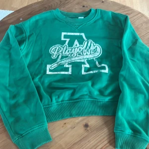 Grön sweatshirt med tryck - Säljer en grön sweatshirt med ett stort vitt tryck på framsidan. Tröjan har en v-ringad hals och ribbade muddar vid ärmslut och nederkant. 