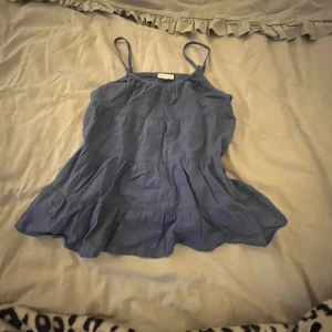 Blå linne från Pomp de Lux - Söt blå linne  från Pomp de Lux med smala axelband och volangdetaljer. Perfekt för varma dagar med sin lätta och luftiga design. Linnet har en avslappnad passform och en liten gulddetalj i form av ett hjärta.