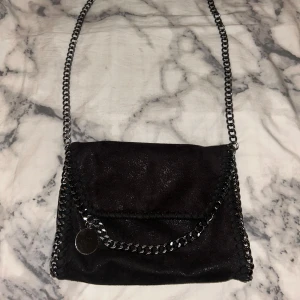 Svart axelväska med kedjedetaljer - Snygg svart axelväska med kedjedetaljer runt kanterna och en kedja som axelrem. Väskan har en rund metallbricka som dekoration. 
