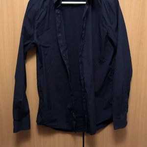 Mörkblå skjorta från COS - Snygg mörkblå skjorta från COS i slim fit. Tillverkad i en blandning av bomull, polyamid och elastan för en bekväm passform. Skjortan har långa ärmar och knappar framtill. Perfekt för en stilren look.